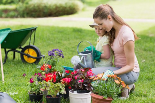 A woman and a young girl are kneeling together on a well-maintained lawn in a garden, engaging in planting or gardening activities. They are surrounded by a variety of colorful potted flowers and plants, including red, pink, purple, and orange blooms, arranged in different sizes and types of containers. In the background, there is a green wheelbarrow with yellow wheels, likely used for garden tools or soil, positioned on the lush, green grass. The garden features a neatly trimmed hedge or shrubbery, providing a natural border, and a pathway can be faintly seen beyond the lawn. The scene is set outdoors on a bright, sunny day, with natural light illuminating the vibrant colors of the plants and the gardening activity. The surroundings suggest a peaceful, landscaped outdoor space suitable for gardening services in the Canary Wharf area, emphasizing the care and attention given to garden maintenance and plant cultivation.