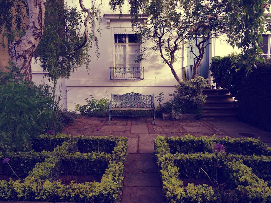 A small urban garden featuring a paved patio area with a decorative metal bench positioned against the white exterior wall of a building, which has a window with a small wrought iron balcony. To the r