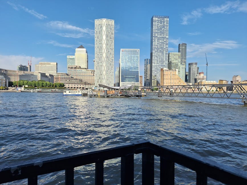 A paved walkway with metal railings on both sides extends along a riverside area of a modern urban landscape. To the right, there is a tall, curved building with large glass windows and balconies, sur