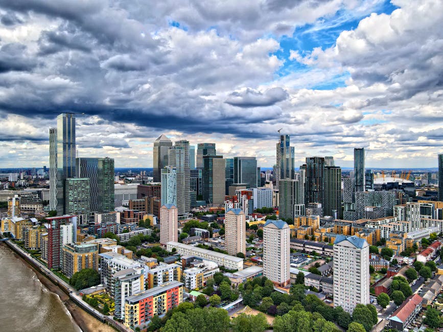A panoramic view of Canary Wharf in London during early evening, showcasing a cluster of modern skyscrapers with glass facades reflecting the cloudy sky. The buildings vary in height and design, inclu